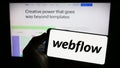 Person holding mobile phone with logo of US web development software company Webflow Inc. on screen in front of web page. Royalty Free Stock Photo