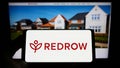 Person holding smartphone with logo of British housebuilding company Redrow plc on screen in front of website.