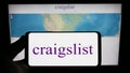 Person holding smartphone with logo of classified advertisements company Craigslist Inc. on screen in front of website.