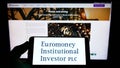 Person holding smartphone with logo of company Euromoney Institutional Investor plc on screen in front of website.