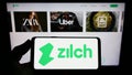 Person holding smartphone with logo of fintech Zilch Technology Limited (PayZilch) on screen in front of website.