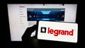 Person holding smartphone with logo of French electronics company Legrand S.A. on screen in front of website.