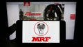 Person holding smartphone with logo of Indian company Madras Rubber Factory (MRF Tyres) on screen in front of website.