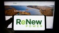 Person holding smartphone with logo of Indian company ReNew Energy Global plc on screen in front of website.