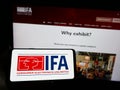 Person holding smartphone with logo of Internationale Funkausstellung Berlin (IFA) on screen in front of website.