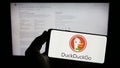 Person holding smartphone with logo of Internet search engine DuckDuckGo (DDG) on screen in front of website.