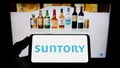 Person holding smartphone with logo of Japanese beverage company Suntory Holdings K.K. on screen in front of website.