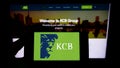 Person holding smartphone with logo of Kenyan financial company KCB Group Limited on screen in front of website.
