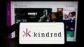 Person holding smartphone with logo of online gambling company Kindred Group plc on screen in front of website.