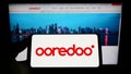 Person holding smartphone with logo of Qatari telecommunications company Ooredoo QSC on screen in front of website.