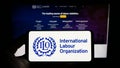 Person holding smartphone with logo of UN agency International Labour Organization (ILO) on screen in front of website.