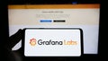 Person holding smartphone with logo of US analytics software company Grafana Labs on screen in front of website.