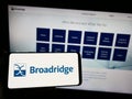 Person holding smartphone with logo of US company Broadridge Financial Solutions Inc. on screen in front of website.