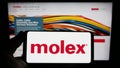 Person holding smartphone with logo of US connector manufacturing company Molex LLC on screen in front of website.