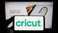 Person holding smartphone with logo of US cutting plotter company Cricut Inc. on screen in front of website.