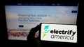 Person holding smartphone with logo of US EV charging company Electrify America LLC on screen in front of website.