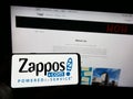 Person holding smartphone with logo of US online shop company Zappos.com LLC on screen in front of website.