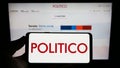 Person holding smartphone with logo of US political newspaper company Politico LLC on screen in front of website.