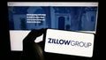 Person holding smartphone with logo of US real estate company Zillow Group Inc. on screen in front of website.