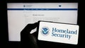 Person holding smartphone with seal of US Department of Homeland Security (DHS) on screen in front of website.