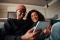 Stylish adult couple, multi-ethnic, laughing at tablet on sofa in modern apartment