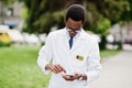 Stylish african american doctor with stethoscope and lab coat, at glasses posed outdoor and using his mobile phone Royalty Free Stock Photo