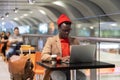 Stylish African American traveler millennial man in beige coat and red hat resting, sitting at cafe table in airport terminal, Royalty Free Stock Photo