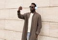 stylish african man taking selfie picture by smartphone in wireless headphones listening to music wearing brown knitted cardigan Royalty Free Stock Photo