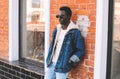 Stylish african man wearing jeans jacket, backpack posing on city street, brick wall Royalty Free Stock Photo