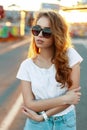 Stylish american redhead young hipster woman in trendy sunglasses in a t-shirt in vintage jeans posing Royalty Free Stock Photo