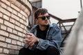 Stylish American young man hipster in trendy blue denim clothes in black sunglasses sits on a vintage staircase near brick wall Royalty Free Stock Photo