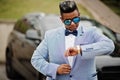 Stylish arabian man in jacket, bow tie and sunglasses against black suv car. Arab rich businessman looking on watches Royalty Free Stock Photo