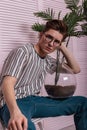 Stylish attractive young male hipster in a vintage striped t-shirt in trendy glasses with a green exotic plant in a glass pot sits
