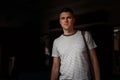 Stylish attractive young man in a fashionable striped t-shirt with a vintage leather bag is standing in a dark room.