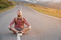 Stylish barefoot bearded male hitchhiker traveler in a hat and with a backpack sits on a suburban asphalt road in the