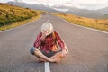 Stylish barefoot bearded male hitchhiker traveler in a hat and with a backpack sits on a suburban asphalt road in the