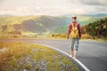 Stylish barefoot bearded male hitchhiker traveler in a hat and with a backpack walks along a country road in the