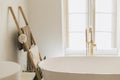 Stylish bathroom interior. Modern bathtub with golden faucet from floor, wooden ladder with towels and big window, boho bathroom Royalty Free Stock Photo