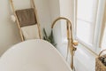 Stylish bathroom interior. Modern bathtub with golden faucet from floor, wooden ladder with towels and big window, boho bathroom Royalty Free Stock Photo