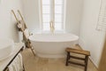 Stylish bathroom interior. Modern bathtub with golden faucet from floor, wooden ladder with towels, rustic bench and big window, Royalty Free Stock Photo