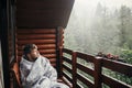 stylish bearded man relaxing on wooden porch among forest in rainy mountains. hipster guy resting in blanket, sitting in