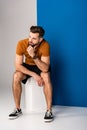 Stylish bearded man in shorts and brown polo sitting on white cube on grey and blue