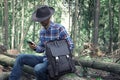 Stylish bearded young man in felt hat, plaid shirt and with trendy leather backpack using his smartphone while sitting on a fallen Royalty Free Stock Photo