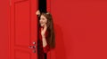 Stylish, beautiful teen girl in trendy red sparkling dress peeking out red door and waving to friends. Party, meeting Royalty Free Stock Photo