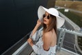 Stylish beautiful young girl in trendy summer clothes with a hat, glasses and a fashion striped top stands near a modern building Royalty Free Stock Photo