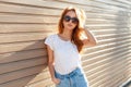 Stylish beautiful young model woman with sunglasses in a white T-shirt and blue vintage jeans near a wooden wall at sunset. Royalty Free Stock Photo