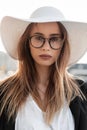 Stylish beautiful young woman in glasses in a fashionable hat with a black jacket and white T-shirt on the street. Young Royalty Free Stock Photo