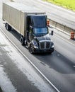 Stylish black big rig day cab semi truck with roof spoiler transporting cargo in container running on the straight divided highway