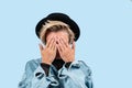 Stylish blond woman closes eyes with her hands in jeans jacket and hat on blue background Royalty Free Stock Photo