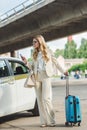 stylish blonde woman in eyeglasses using smartphone and holding suitcase while standing Royalty Free Stock Photo